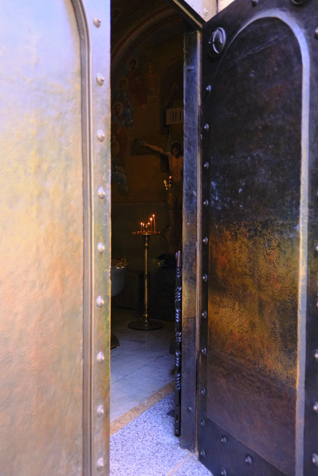 An open studded metal door frames a candle stand and frescoes inside a church.
