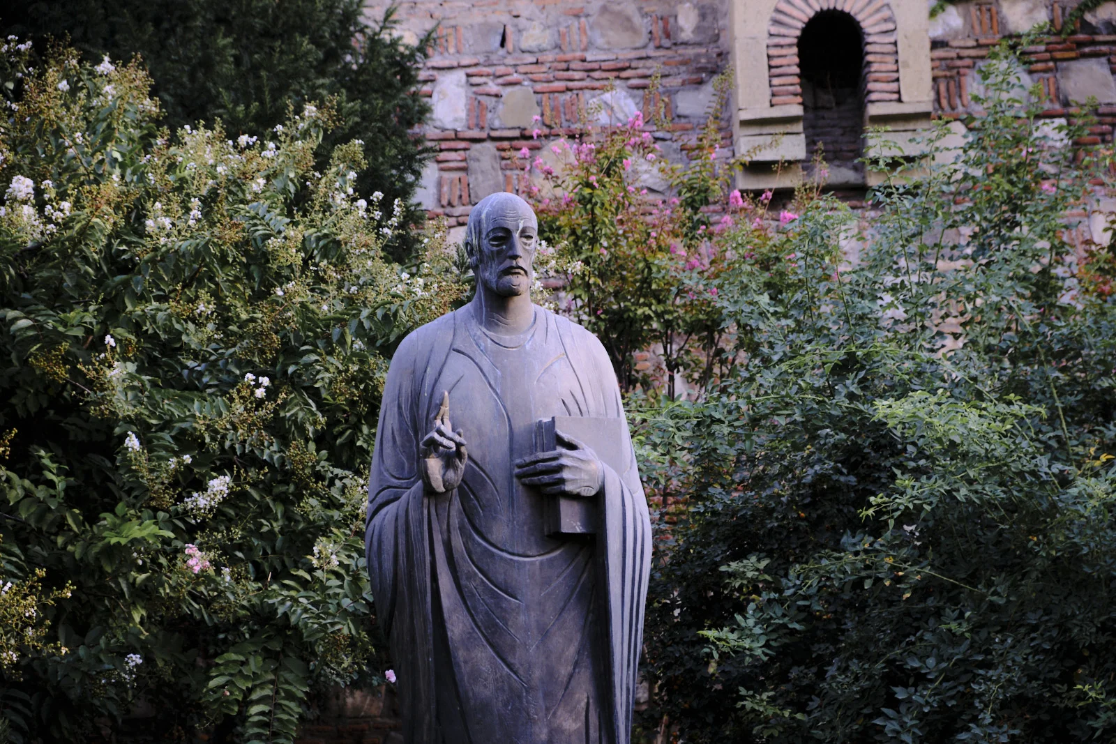 A bronze statue of a robed man holding a book stands among flowering bushes before a brick wall.