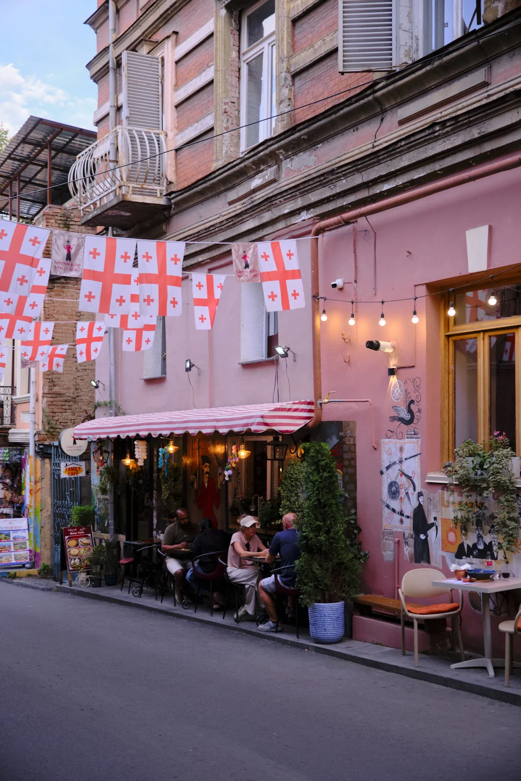 Georgian flags hang over an outdoor café where people sit beneath a striped awning.