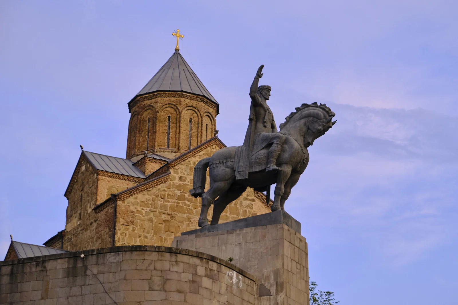 An equestrian statue with a raised arm stands before a stone church with a conical dome and a cross.