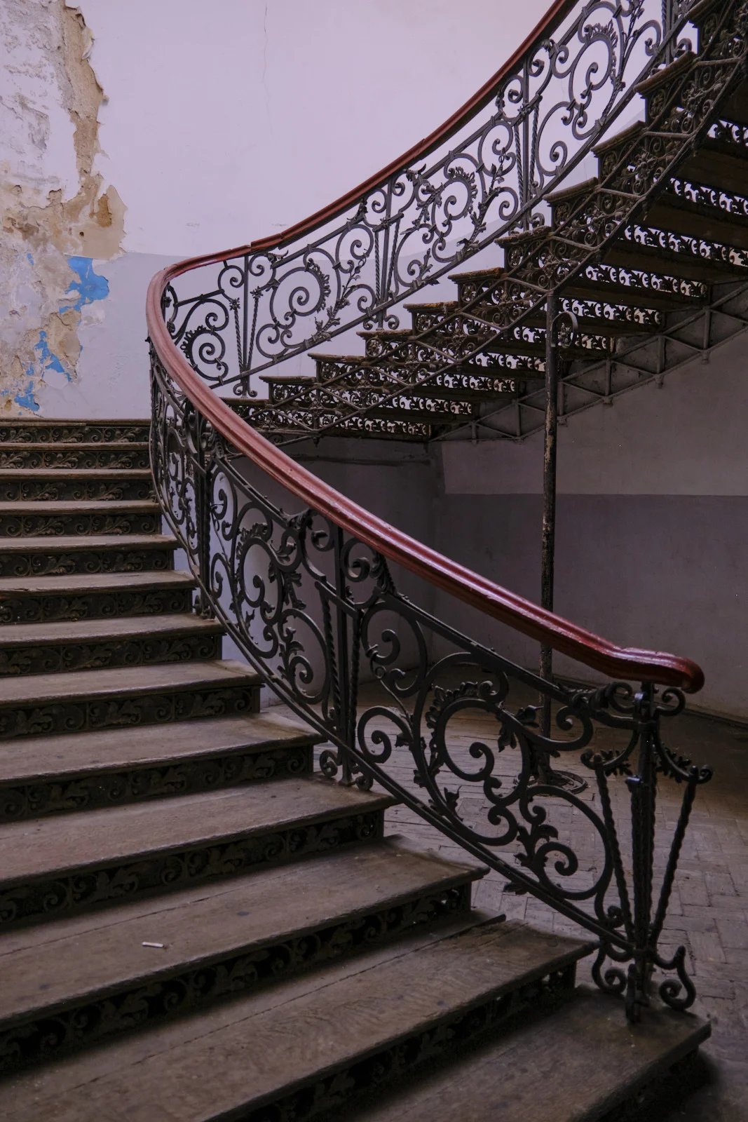 A curving wooden staircase with ornate iron railings and worn steps fills a peeling stairwell.