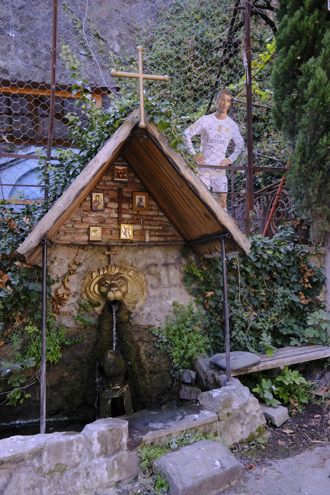 A lion-head spring flows under a wooden shrine with icons and a cross, with a footballer cutout behind a fence.