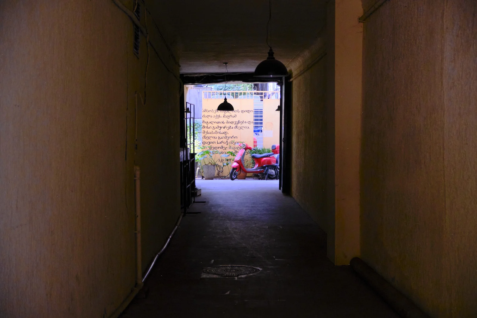 A dim passage frames a red motor scooter parked before a yellow wall covered in Georgian script.