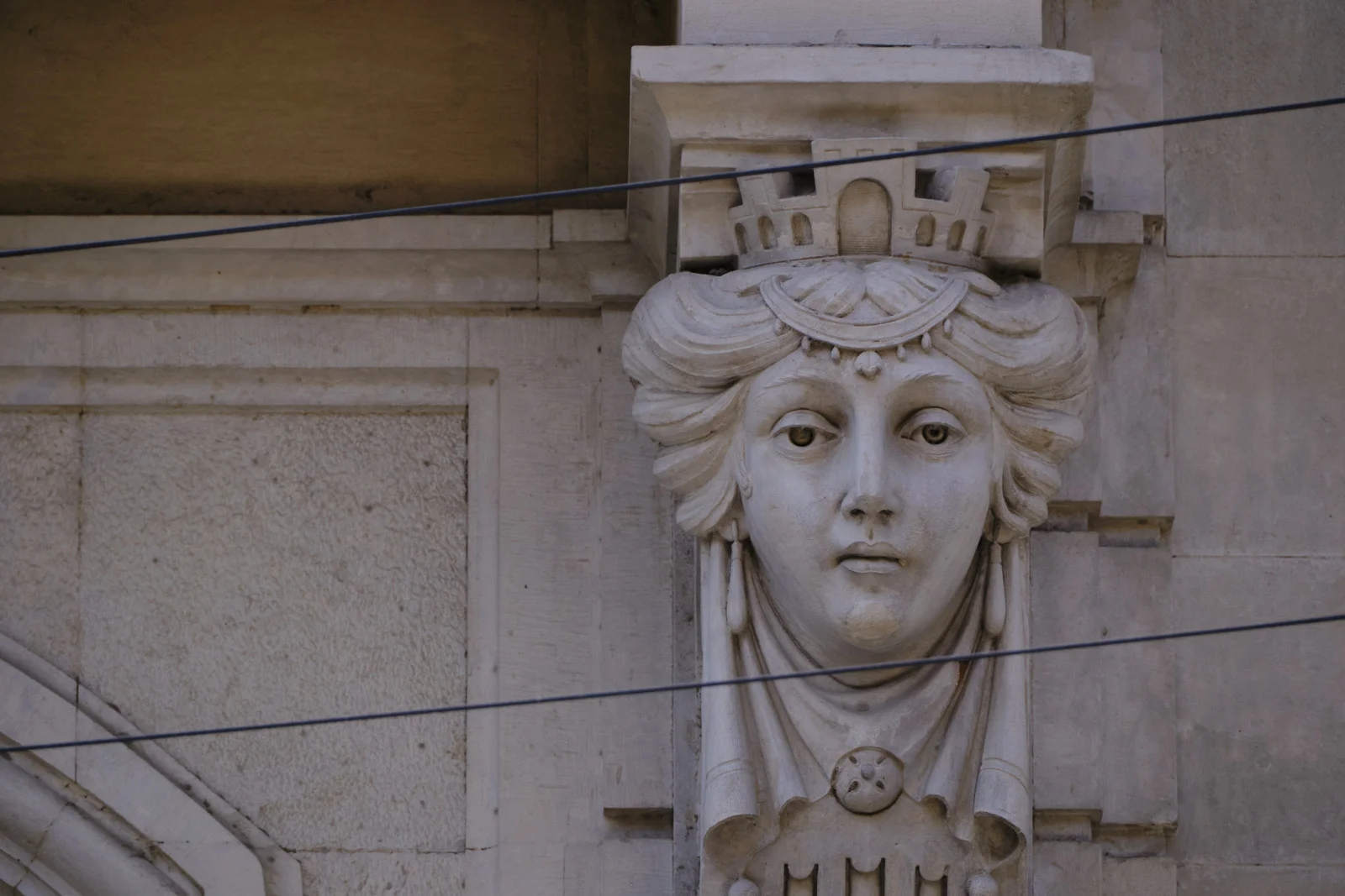 A stone head of a crowned woman used as a building ornament, with wires crossing in front.