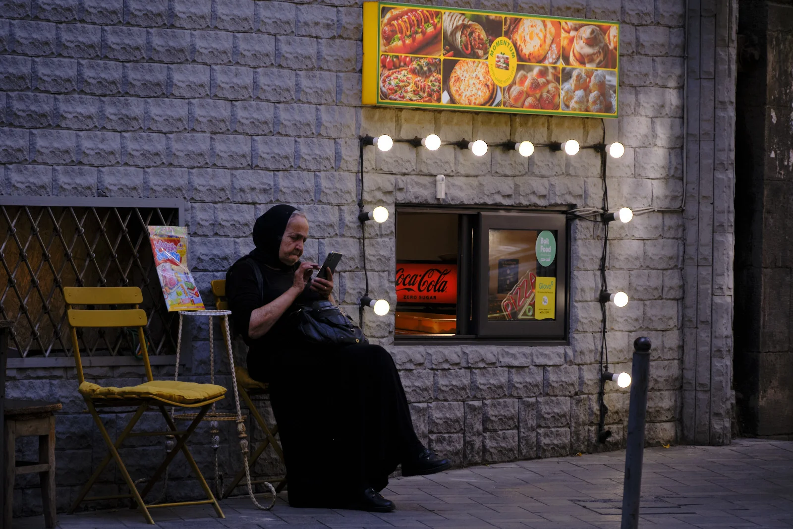 A woman dressed in black sits against a stone wall looking at her phone beneath a lit food-stand window.