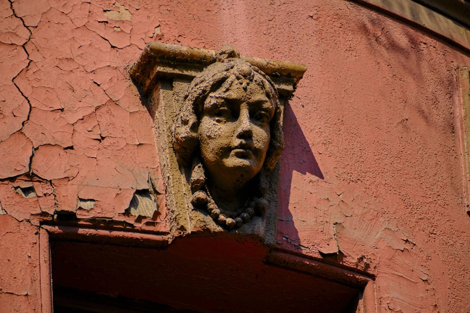 A sculpted woman's face with beads and a leafy headdress is set into cracked pink plaster above a doorway.