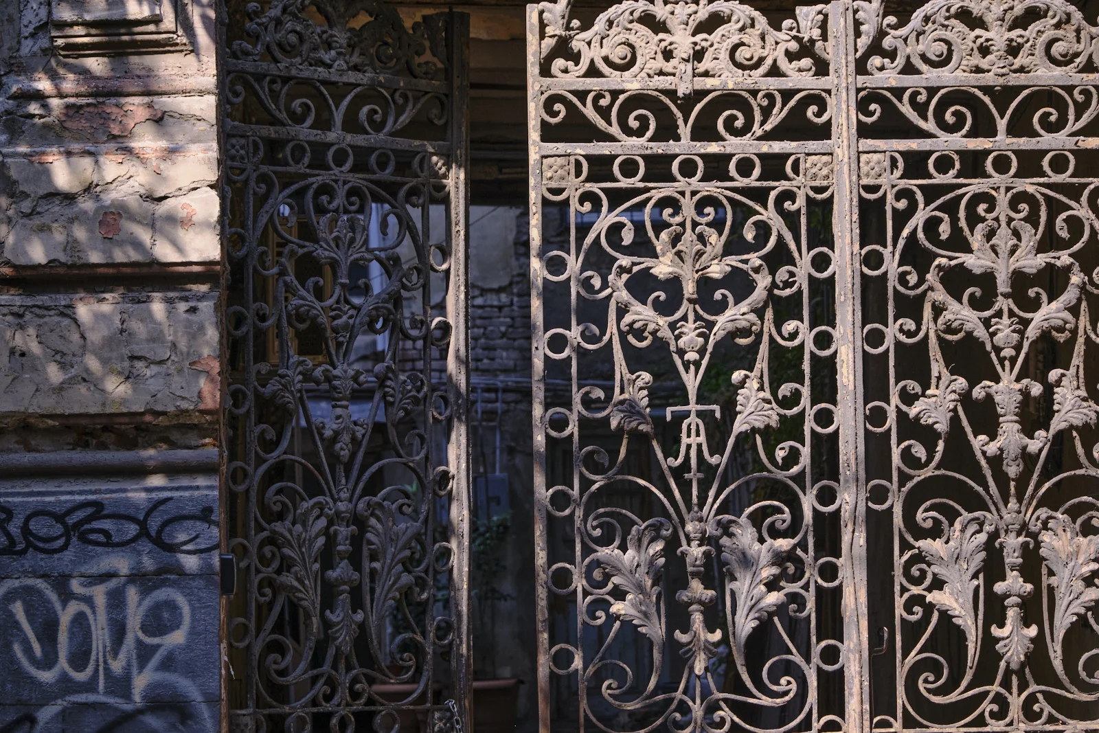 An ornate iron gate standing open against a crumbling brick wall with graffiti.