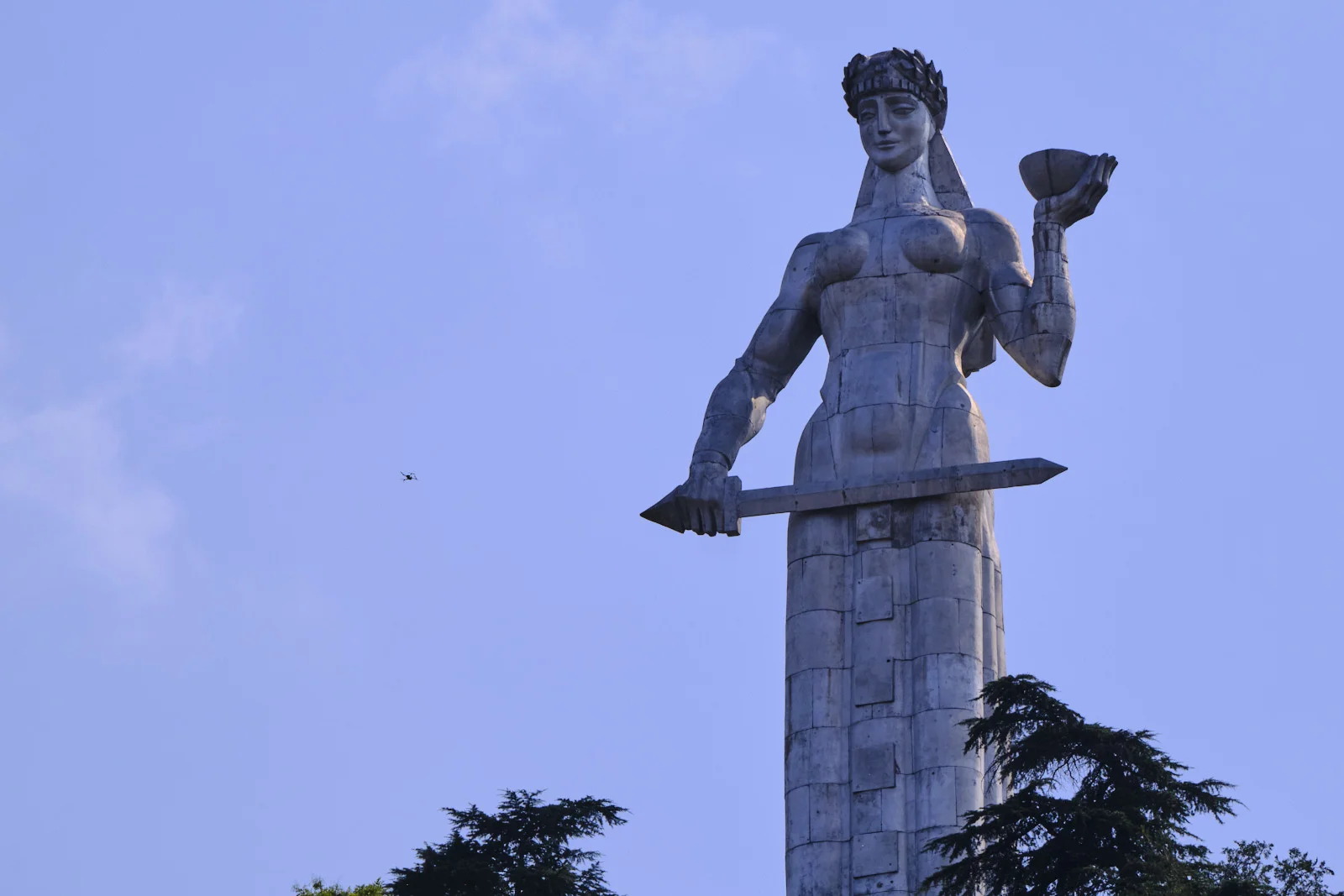 A monumental metal statue of a woman holding a bowl and a sword stands against the sky above trees.