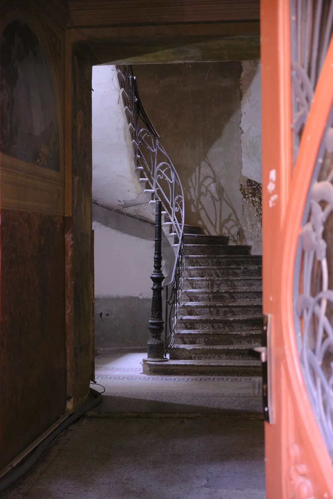 An open orange door frames a worn stone staircase with an ornate iron railing.