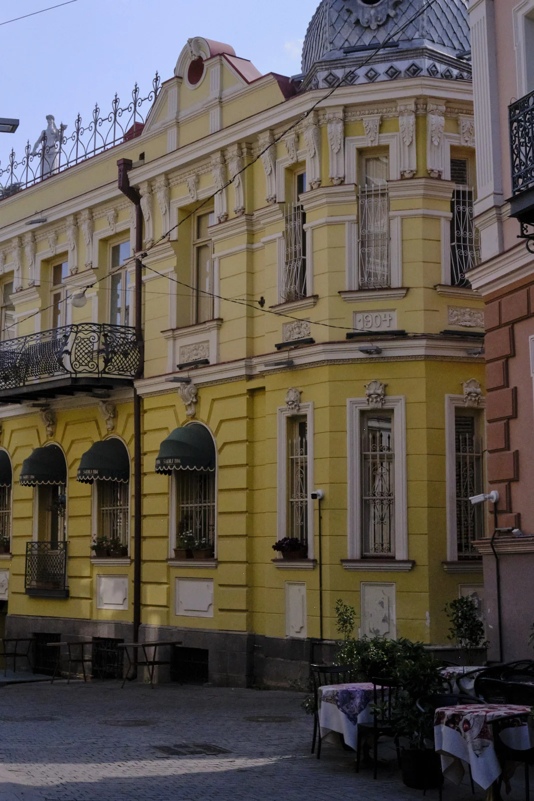 A yellow corner building with green awnings and a silver dome, in a shaded square.