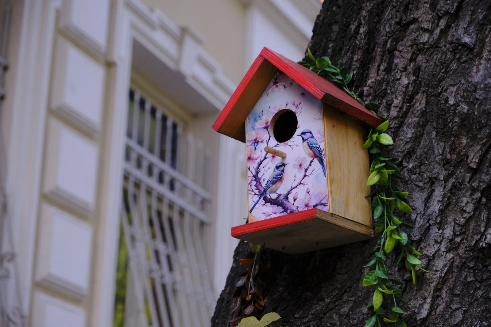 A decorative wooden birdhouse with a red roof and pictured birds is mounted on a tree trunk.