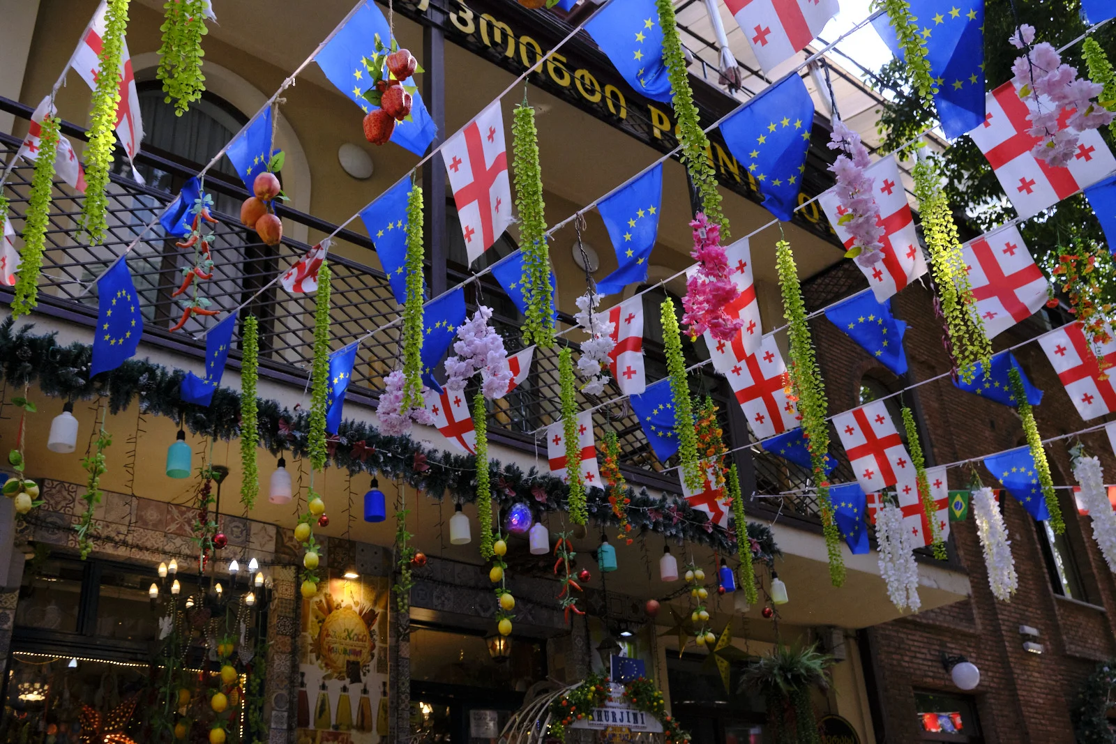 Georgian and European Union flags hung with flowers and fruit under a covered arcade.