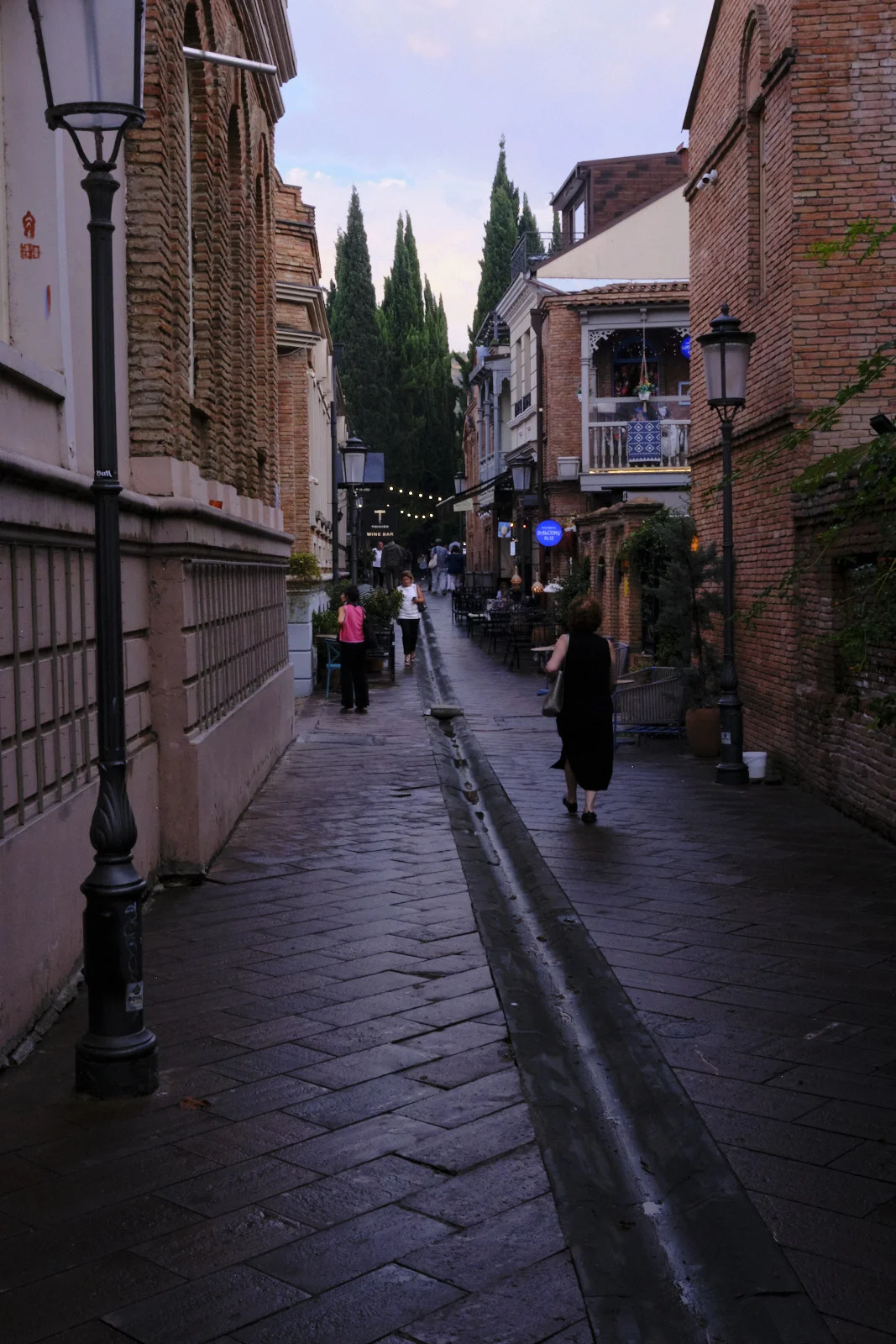A wet brick lane at dusk between brick buildings, with people walking toward lit balconies.