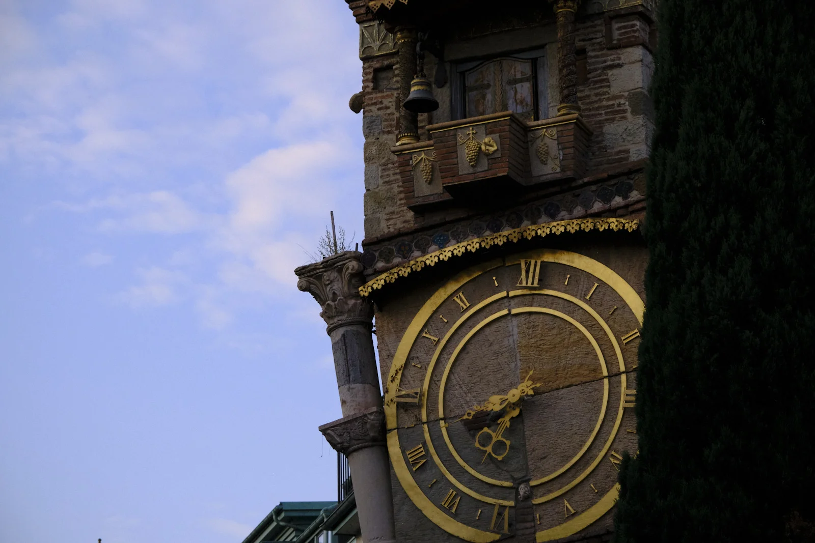 The leaning clock tower with a gold Roman-numeral face, a bell, and a small balcony of grapes.