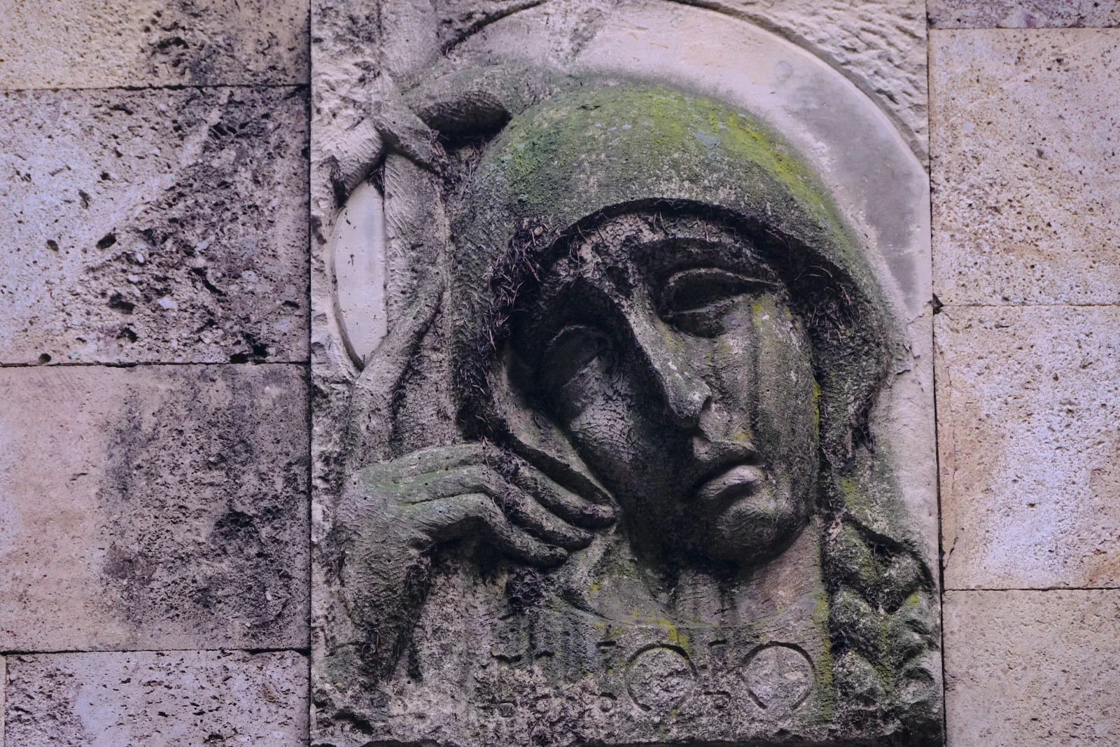 A mossy stone relief of a hooded woman resting her face on her hand is set into a block wall.