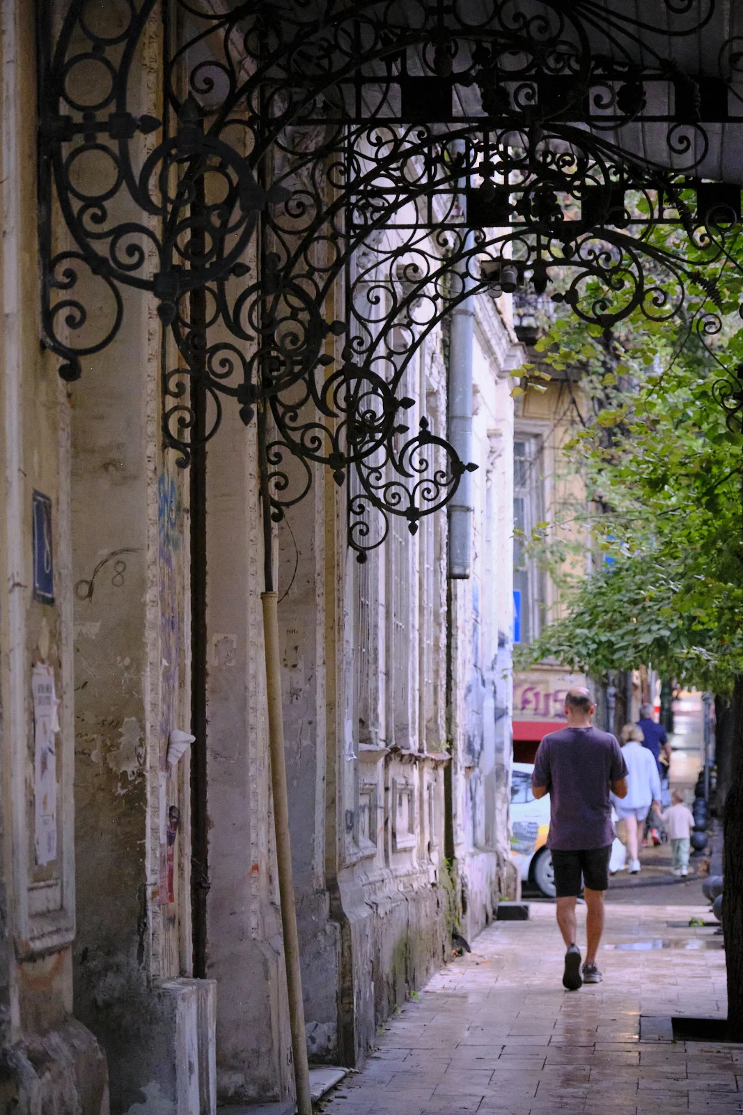 An ornate wrought-iron awning extends over a wet sidewalk as a man walks away along a weathered street.