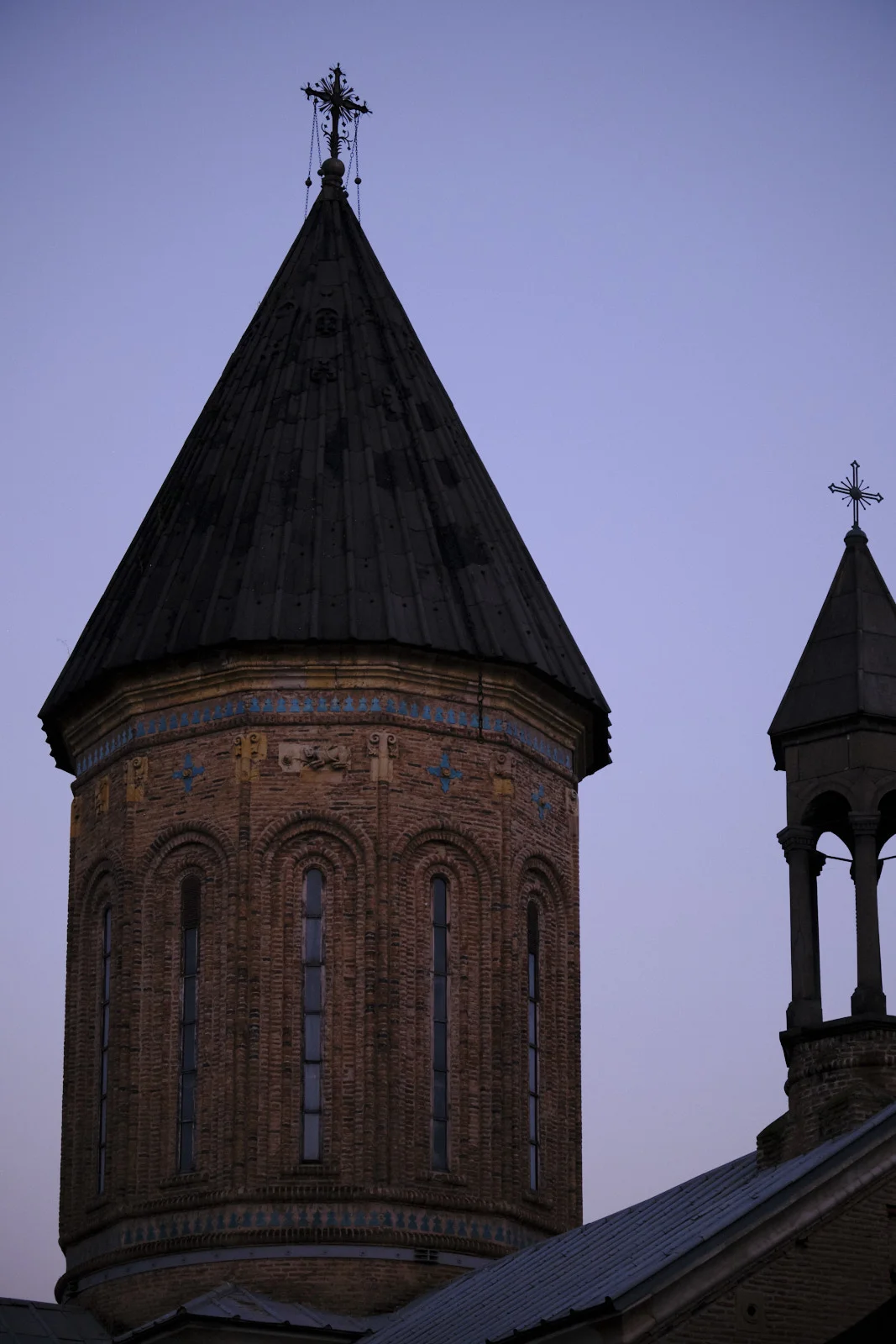 A brick church tower with a dark conical roof and a cross, against a violet evening sky.