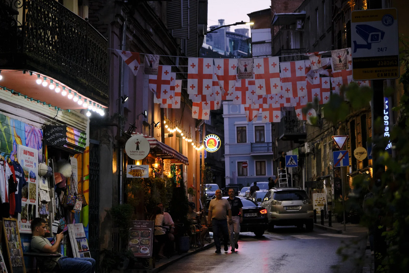 Georgian flags hang across a wet dusk street with pedestrians, a café, and a souvenir stall.