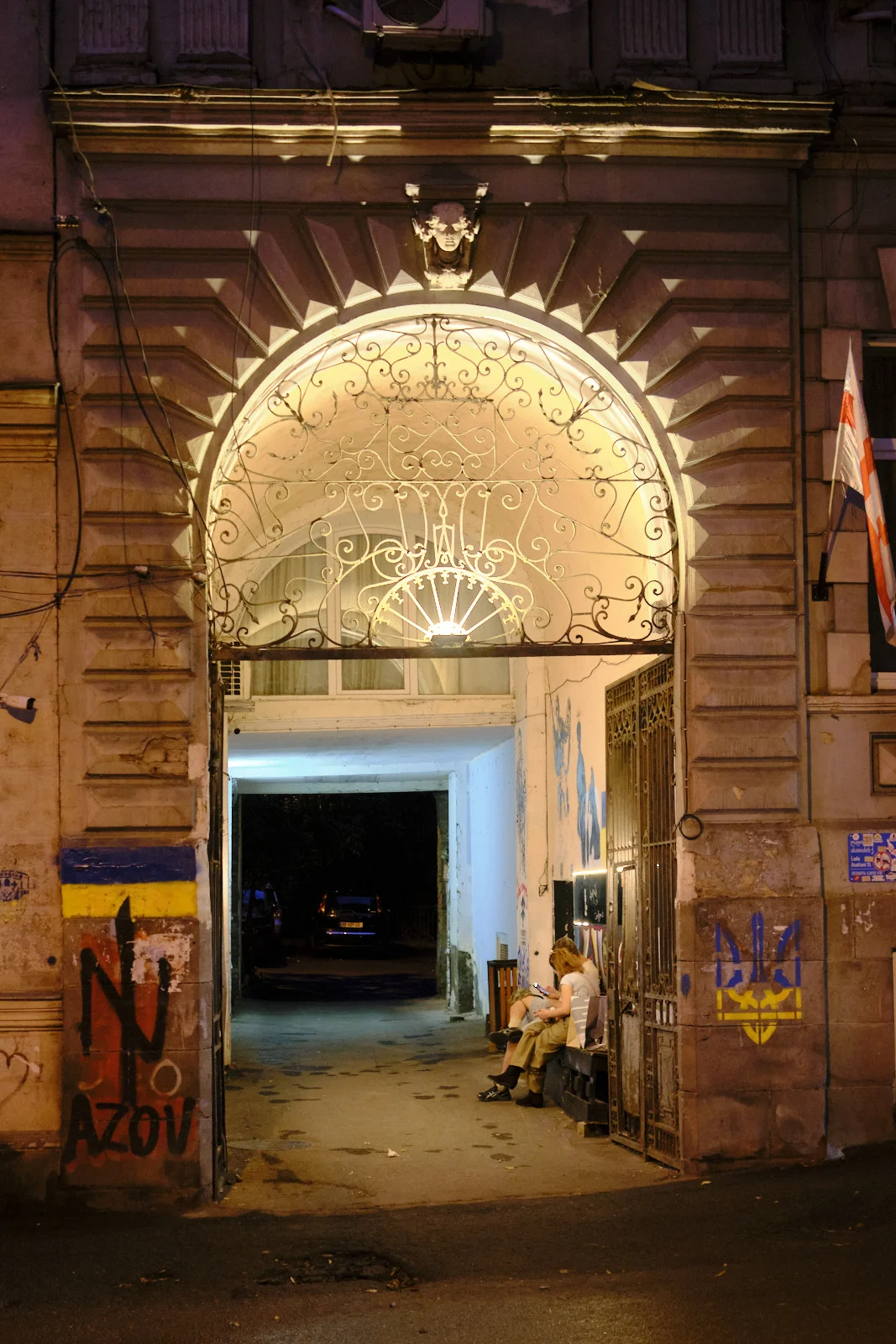 A nighttime stone archway with an ornate iron grille, painted graffiti on the pier, and people sitting in the passage.