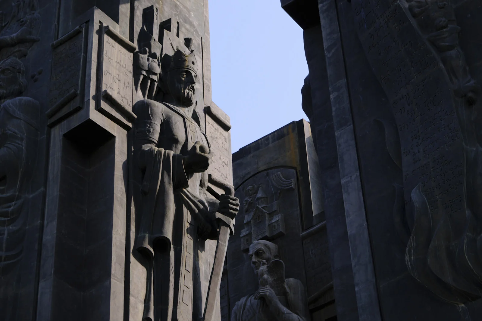 A dark stone relief of a crowned king holding a sword, on the Chronicle of Georgia monument.