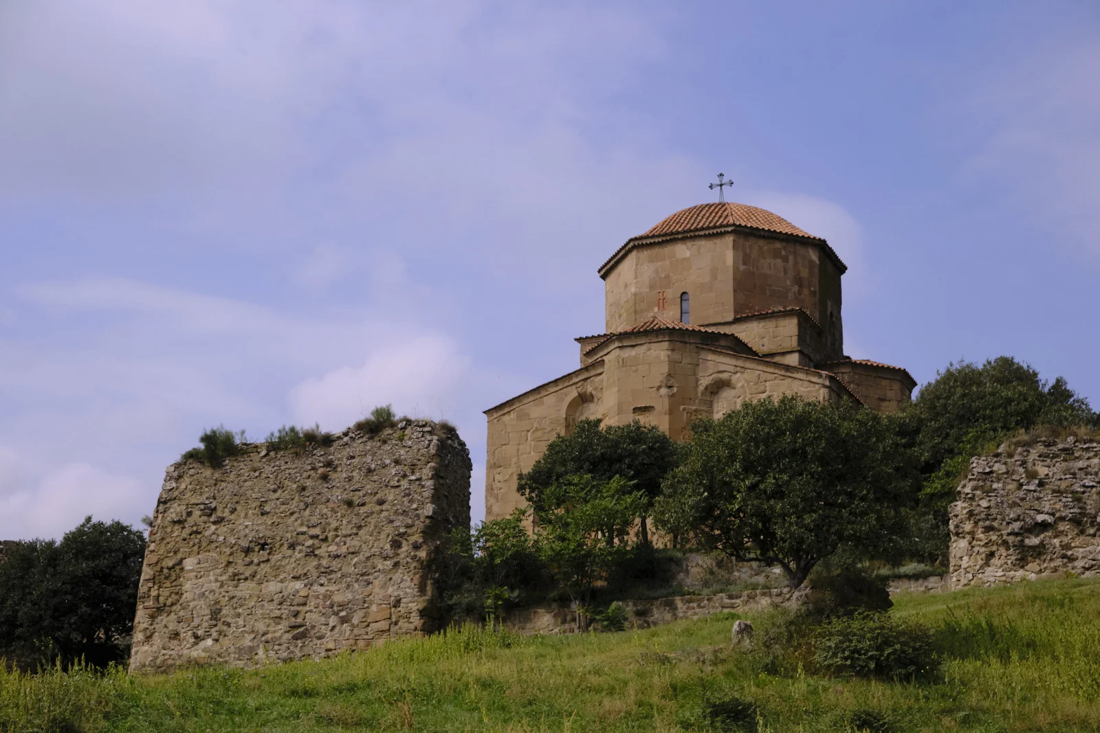 Jvari Monastery on a green hill, a ruined stone wall in the foreground.