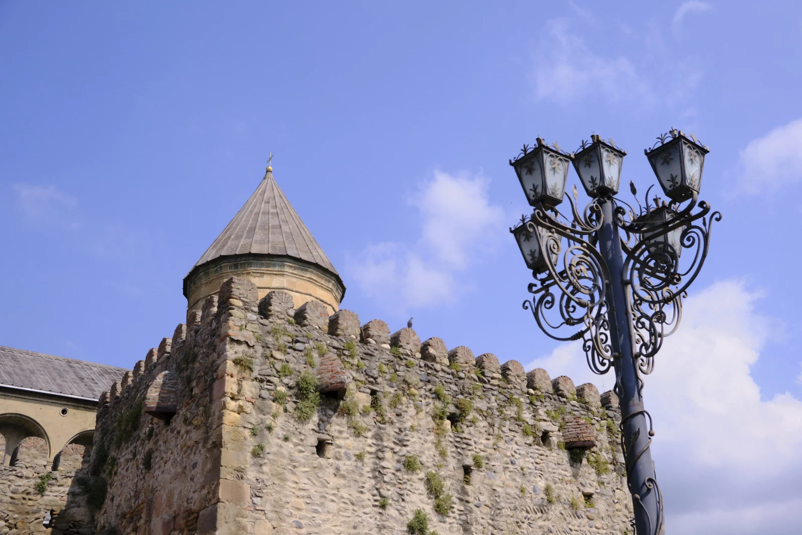 An iron lamp post in front of the crenellated stone wall and conical tower of Svetitskhoveli.