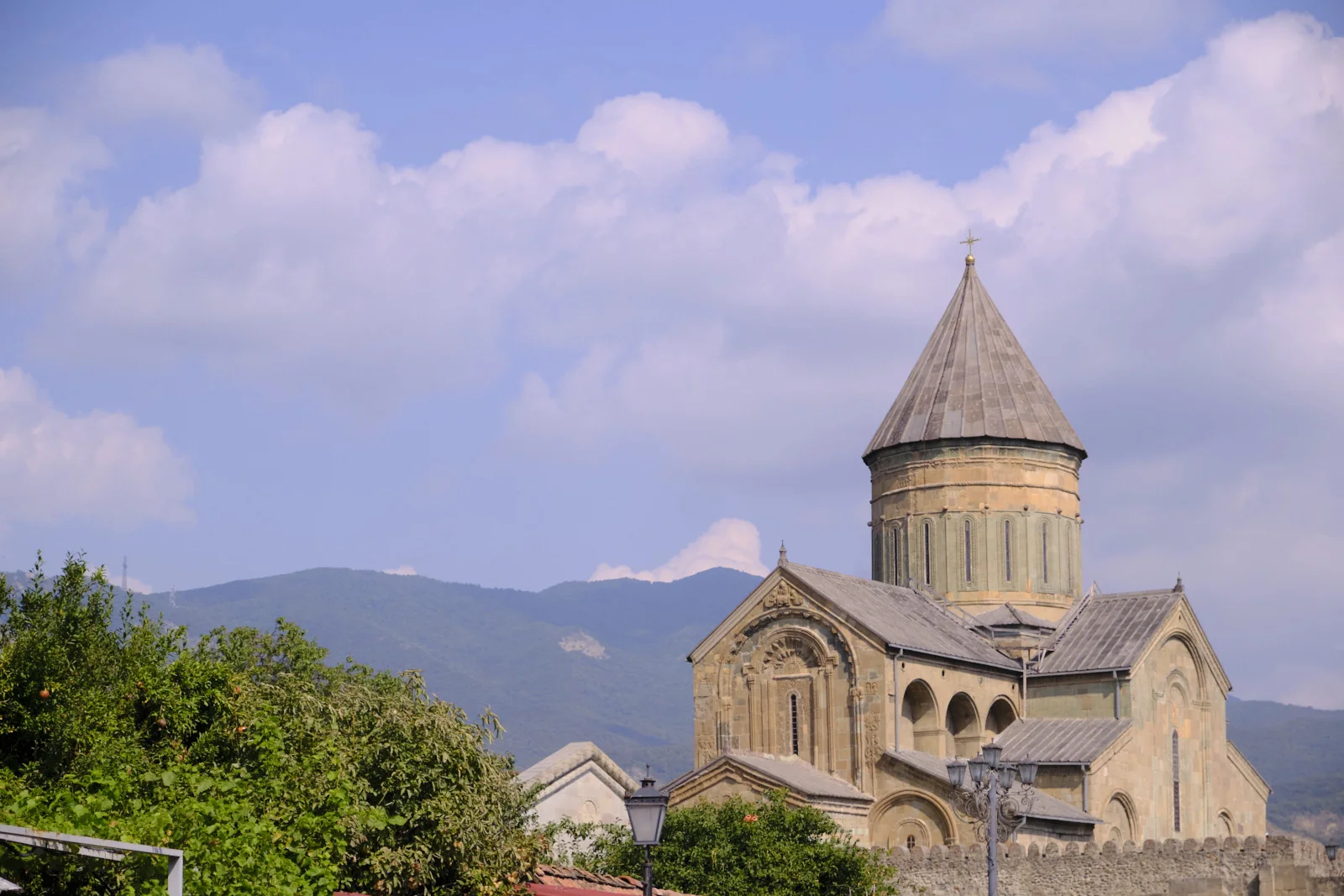 A stone church with a conical roof, trees in front and blue mountains behind.