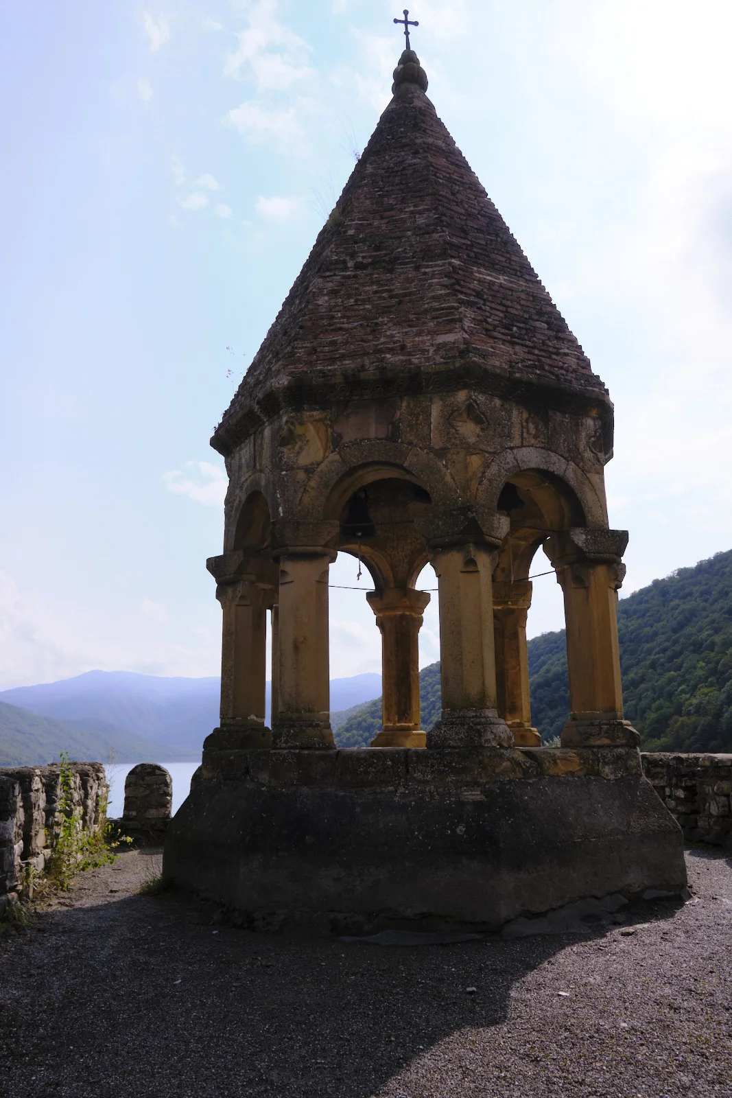 A small open bell tower with a tiled roof, mountains and the reservoir behind it.