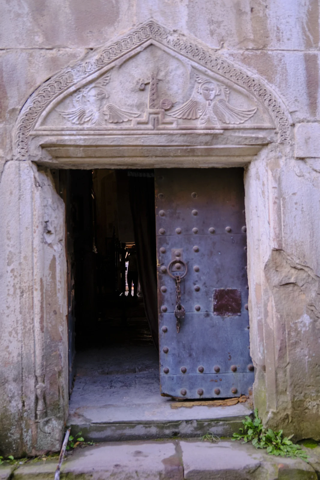 A dark studded door under a carved tympanum of two angels flanking a cross.