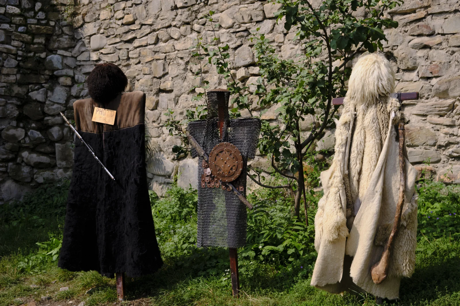 Three costume displays against a stone wall: a dark cloak, chain mail, and a white sheepskin.