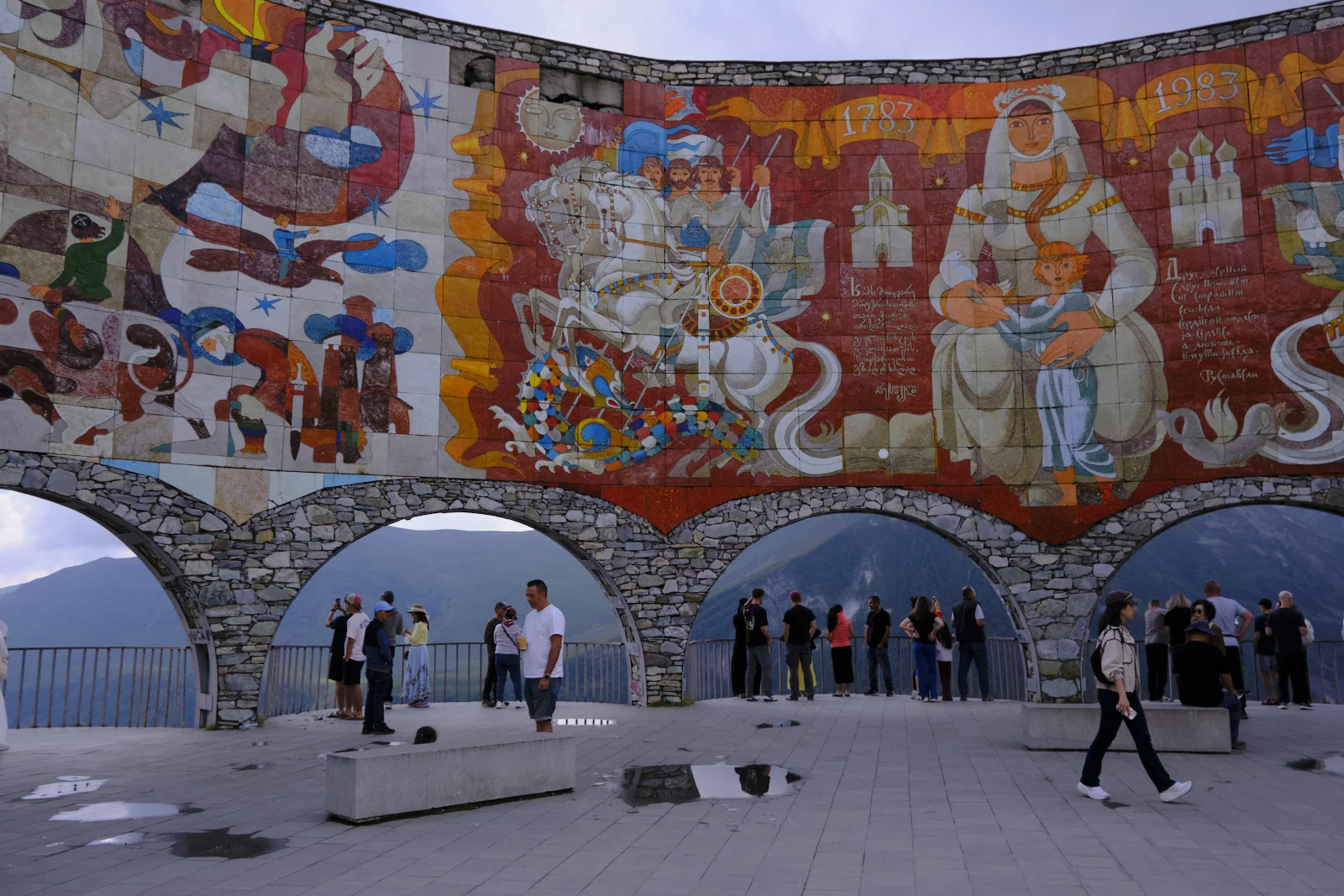 Visitors on a terrace under a huge curved mosaic of Georgian history, dated 1783 and 1983.