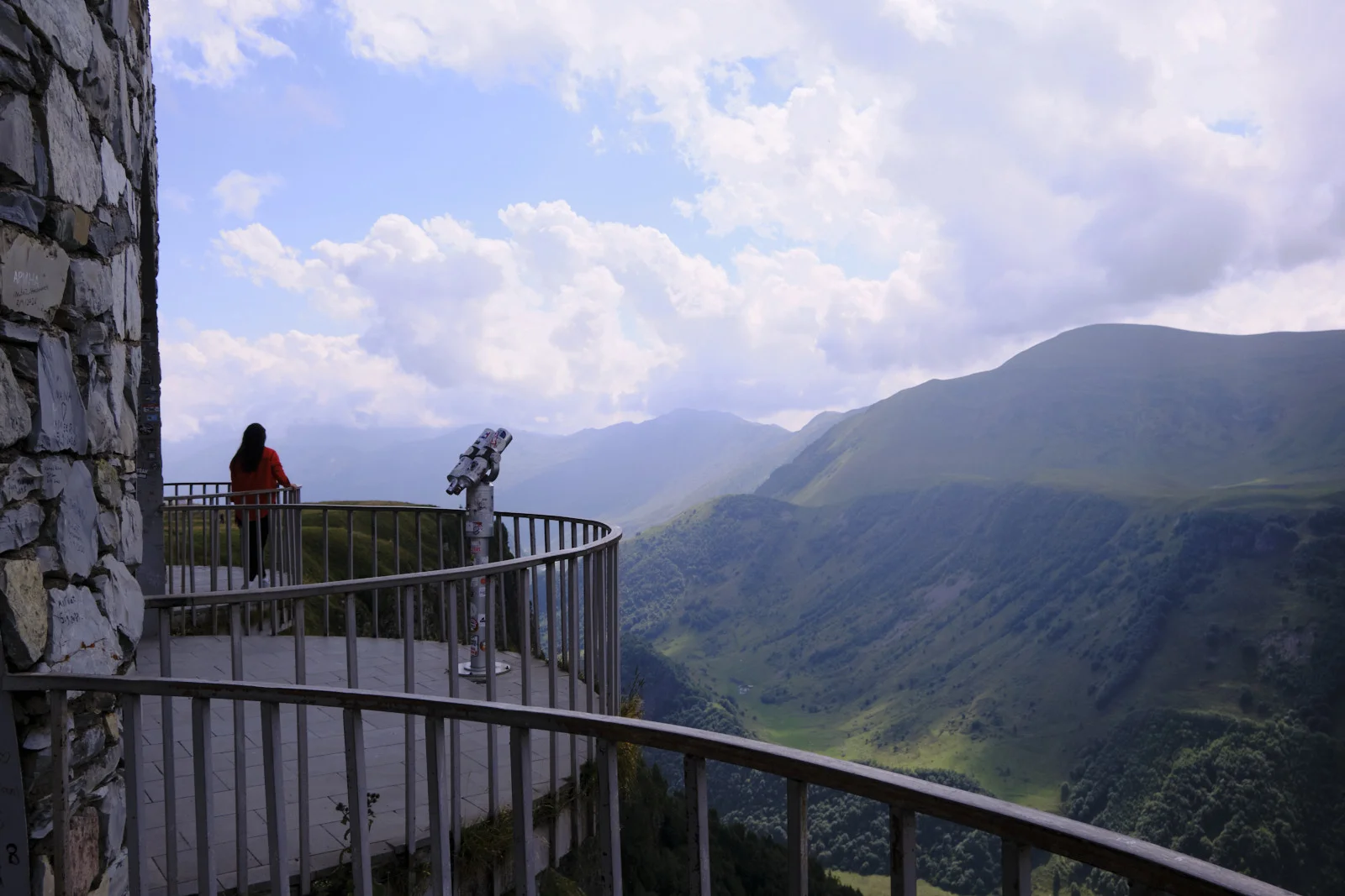 Two people at a curved railing on a stone terrace, looking over a wide green valley.