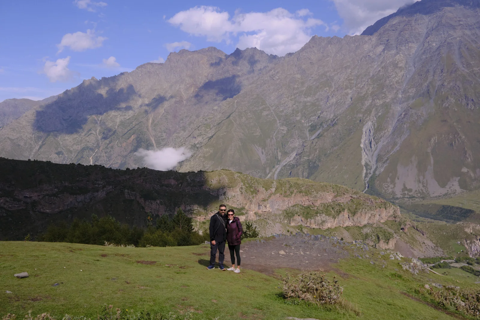 A couple standing on a grassy slope with the mountains of Kazbegi behind them.