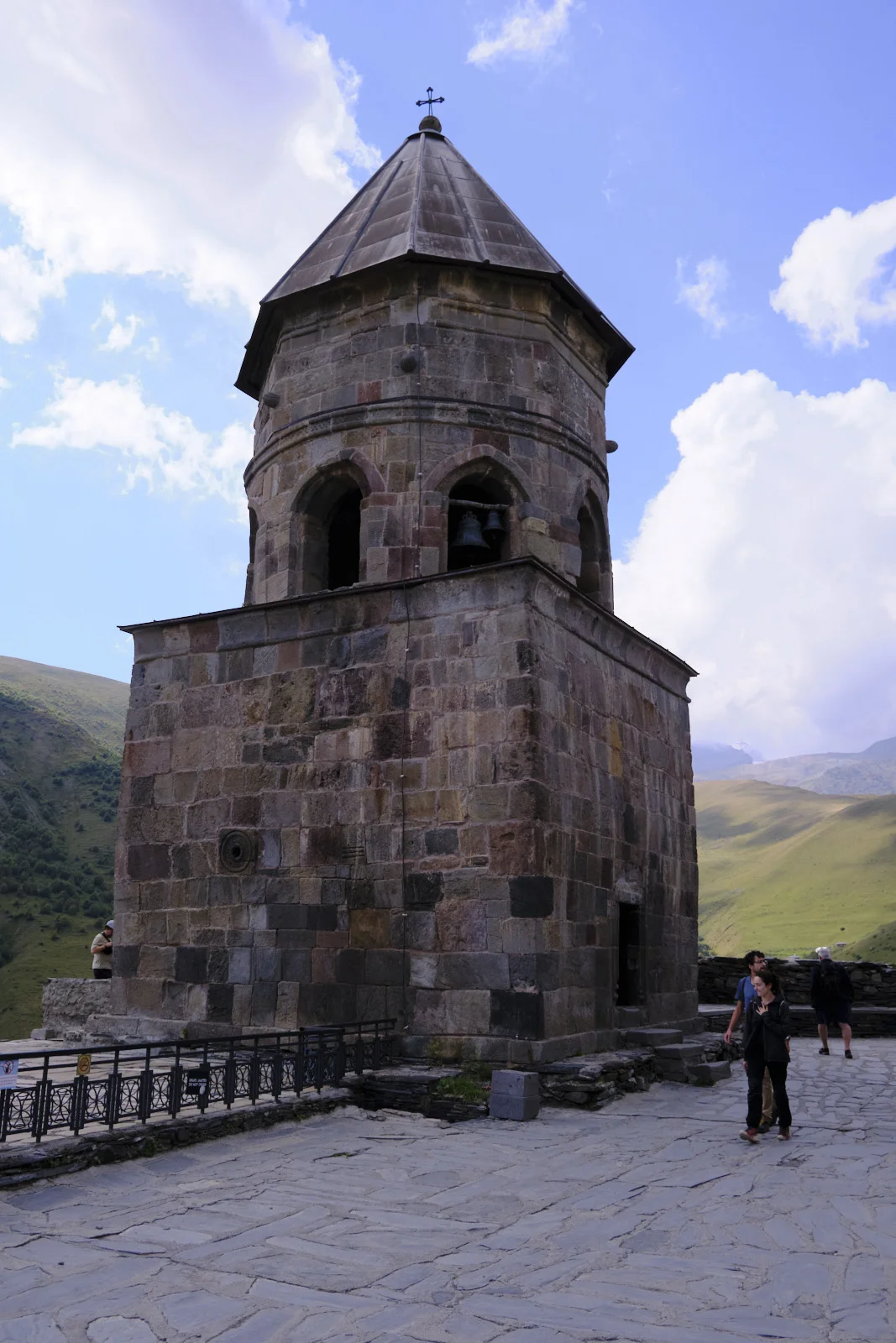 The stone bell tower of Gergeti Trinity, mountains and pasture behind it.