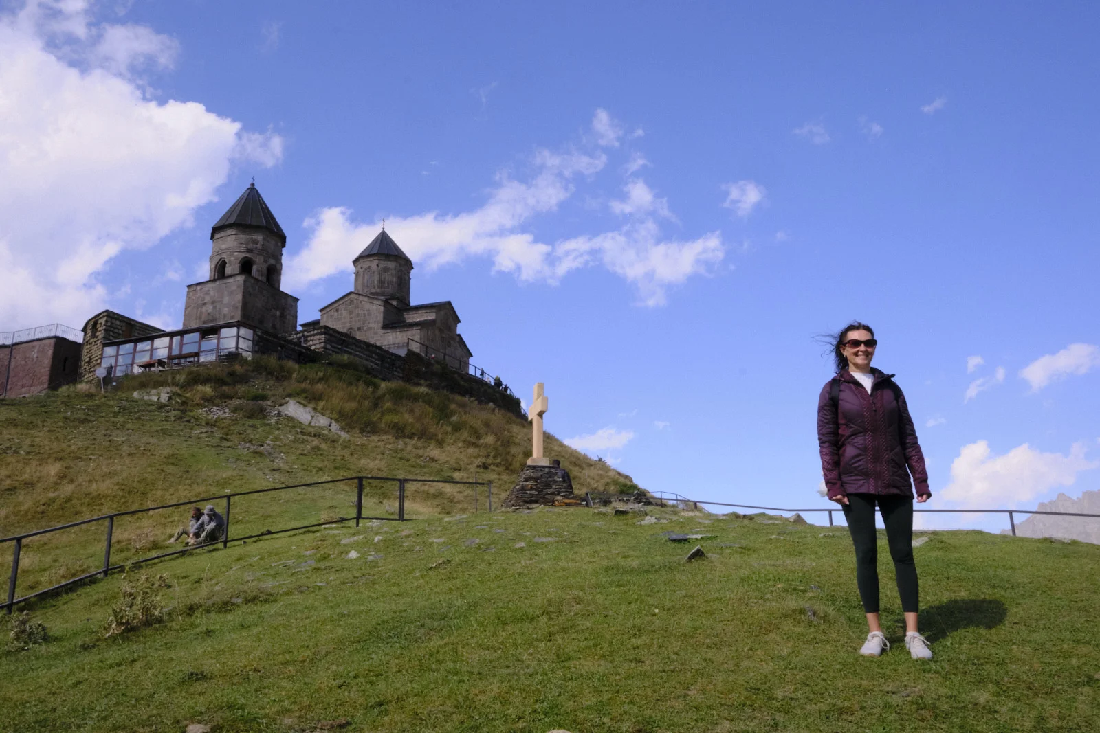 A woman in a purple jacket standing on the grass below Gergeti Trinity Church.