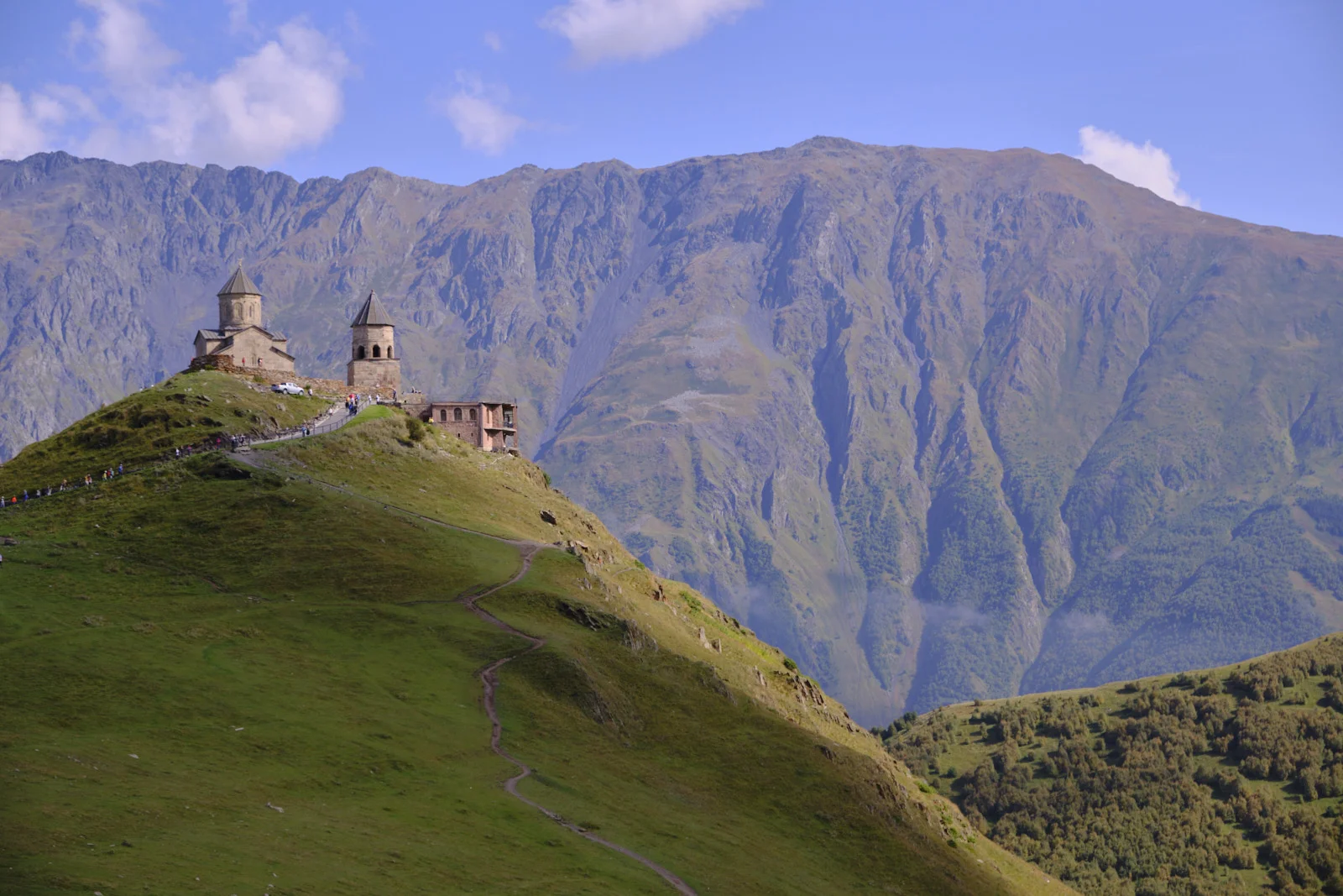 A path up the green hill to Gergeti Trinity, a huge mountain wall behind.
