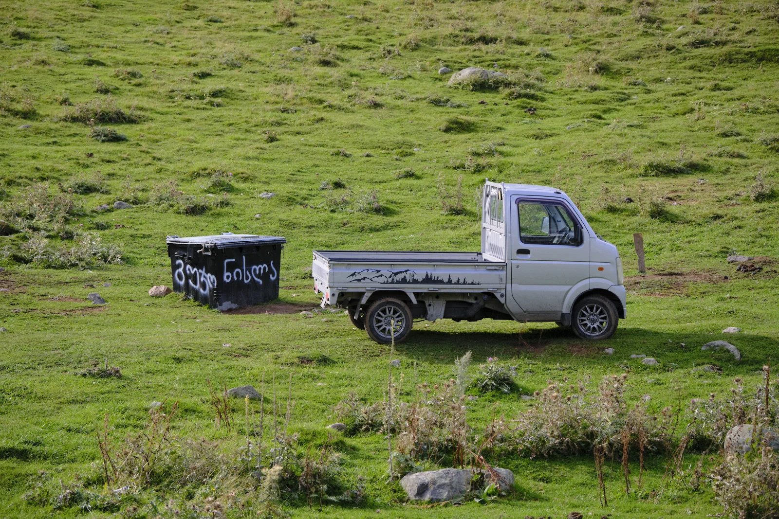 A small white truck and a black dumpster on a green mountainside pasture.