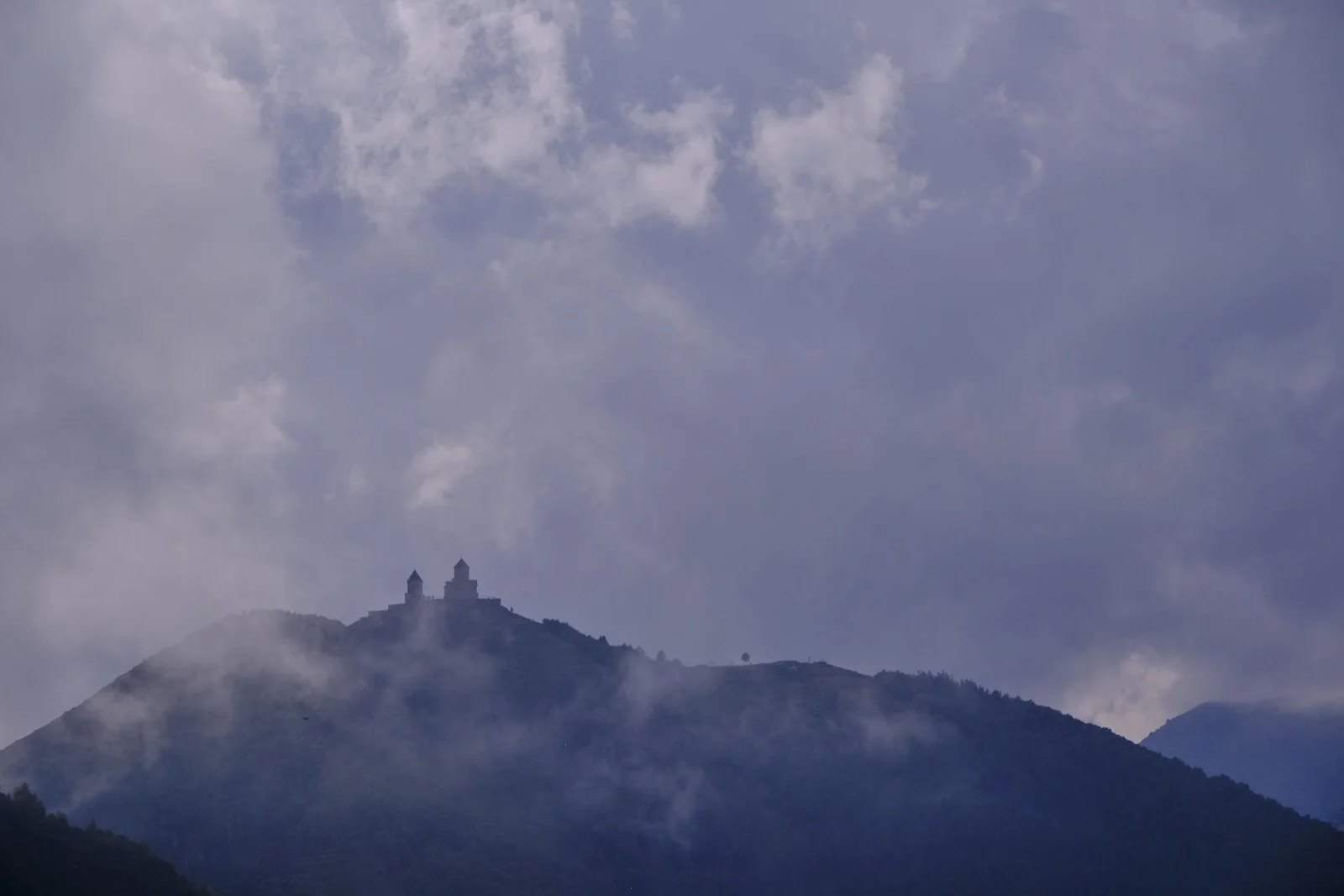 Gergeti Trinity reduced to a small silhouette on a ridge, in heavy mist.