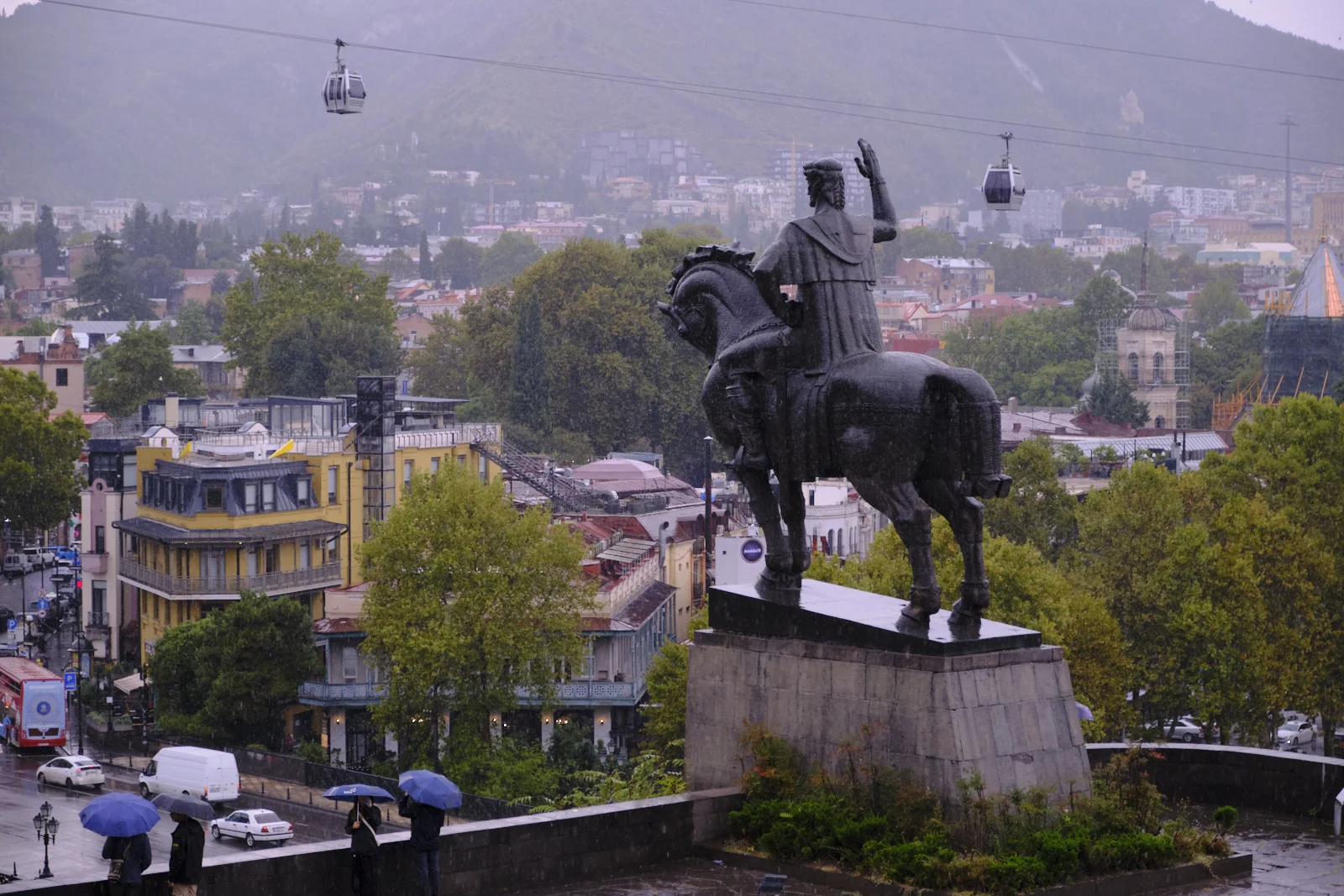 The equestrian statue of Vakhtang Gorgasali in the rain, cable cars overhead and umbrellas in the street.
