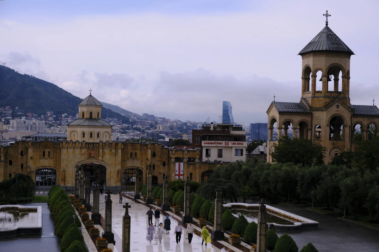 The Holy Trinity Cathedral of Tbilisi, its bell tower and gate, a wet path and the city beyond.