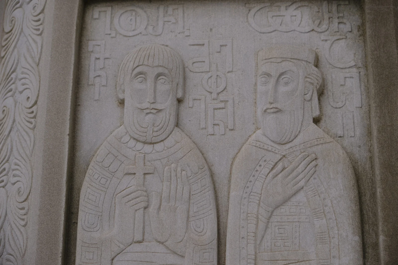 A pale stone relief of two haloed saints, one holding a cross, with Georgian letters above.