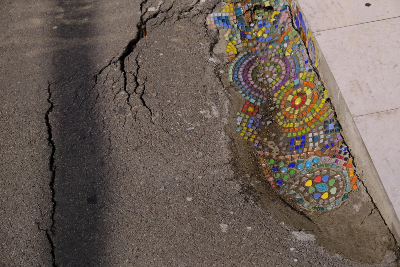 A cracked sidewalk with a strip of colorful mosaic set into the broken edge.