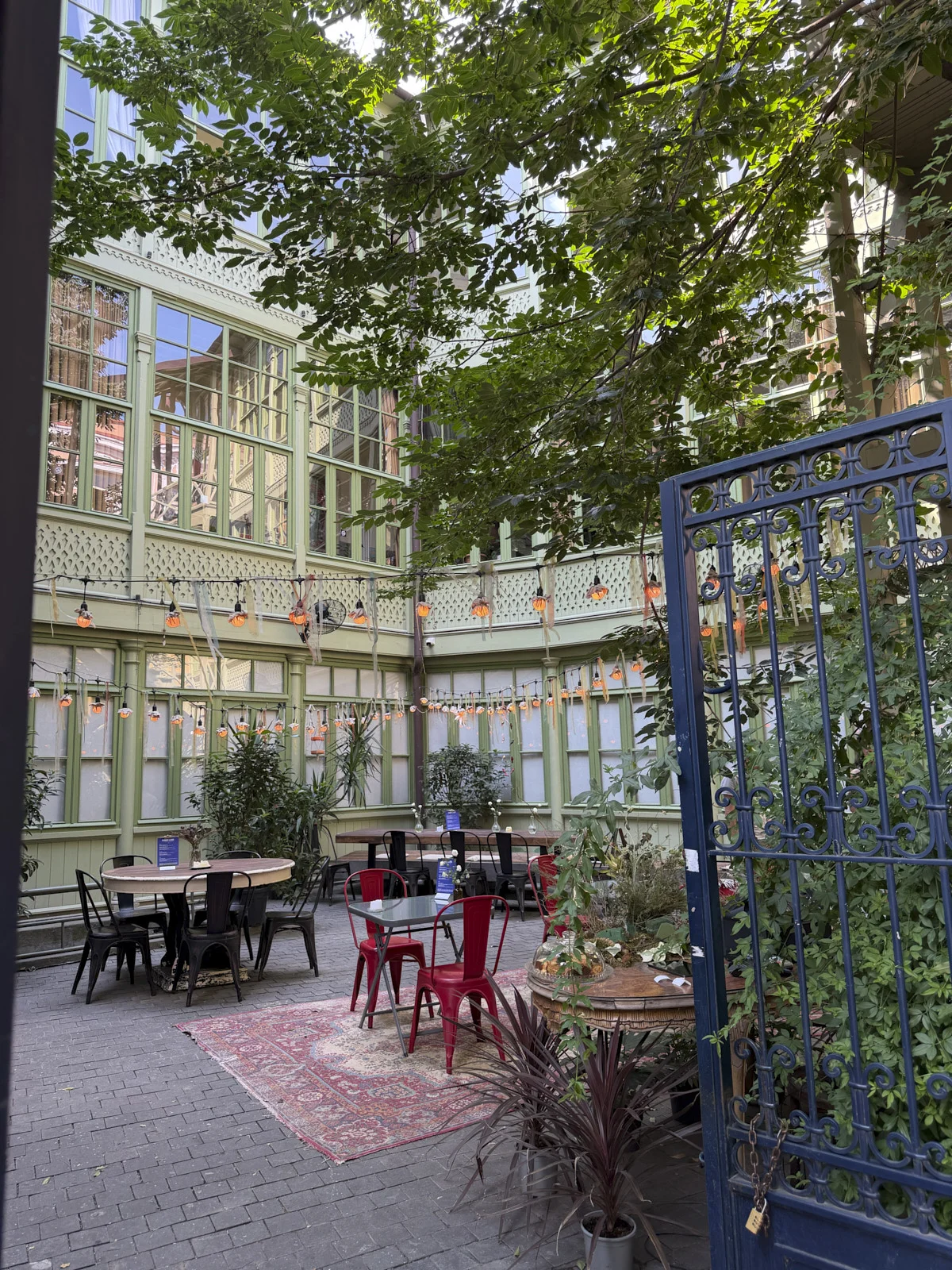 A planted courtyard seen through a blue iron gate, with red chairs and strings of orange lights.