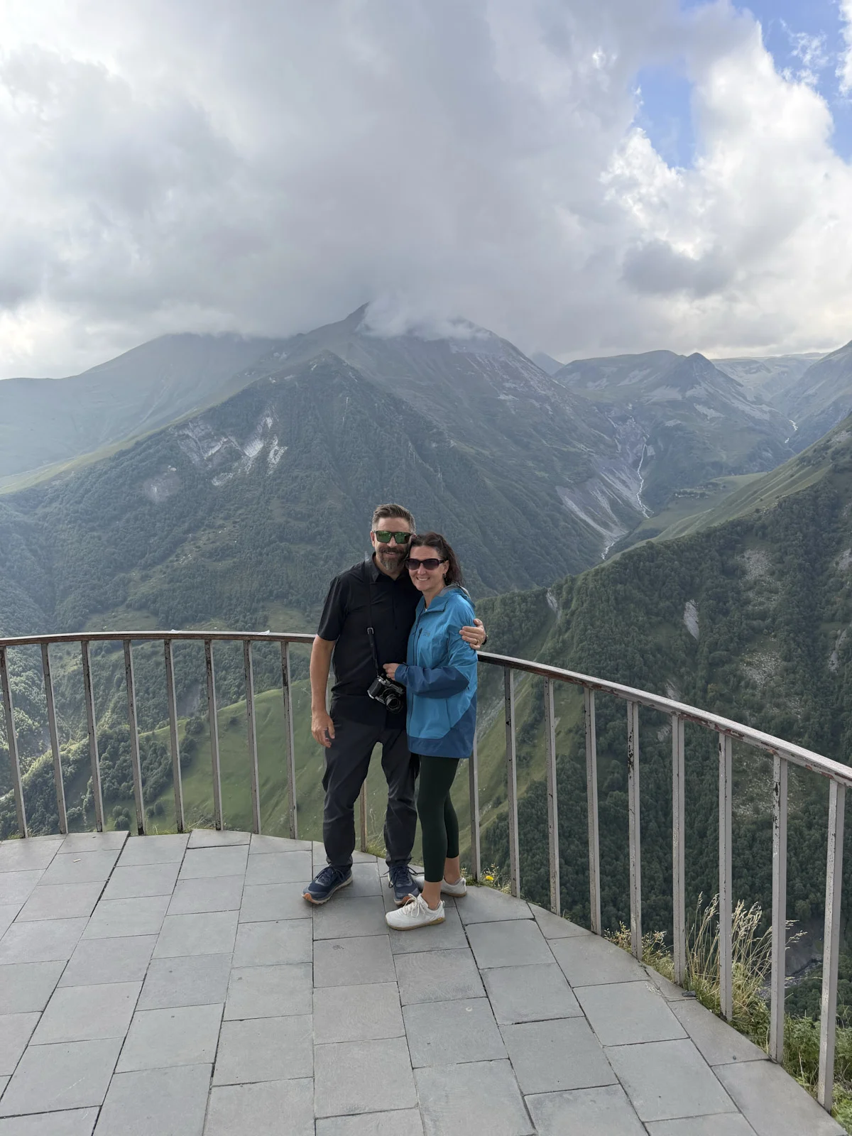 A couple standing at a metal railing on a high overlook, steep green mountains behind them.