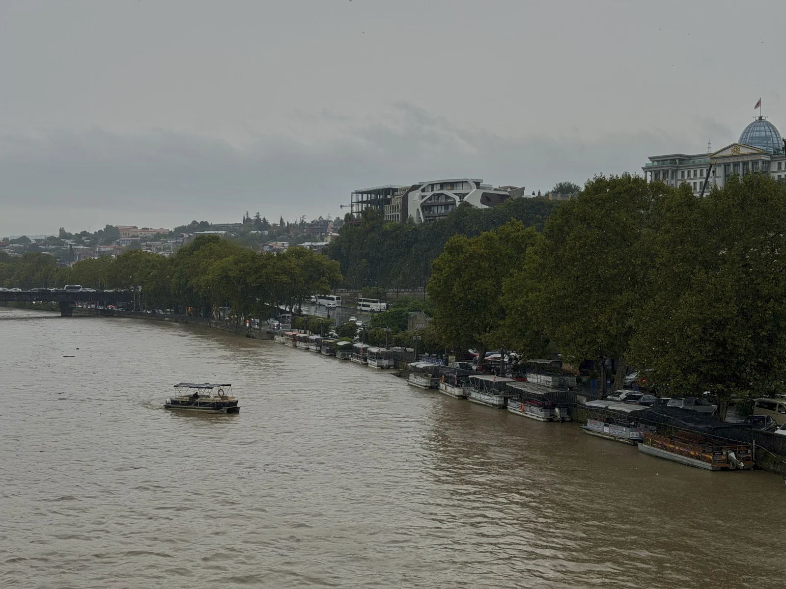 A gray river with a small boat and a row of docked launches, trees and a domed building on the far bank.
