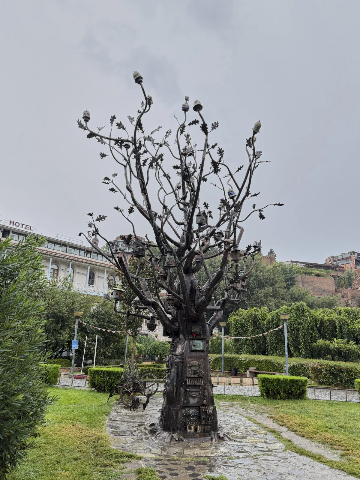 A metal sculpture of a tree with leaves and birds, in a small paved garden.