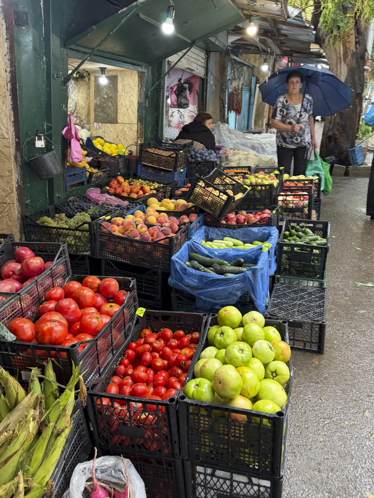 Market crates of tomatoes, apples, and corn, a woman with a blue umbrella beside them.