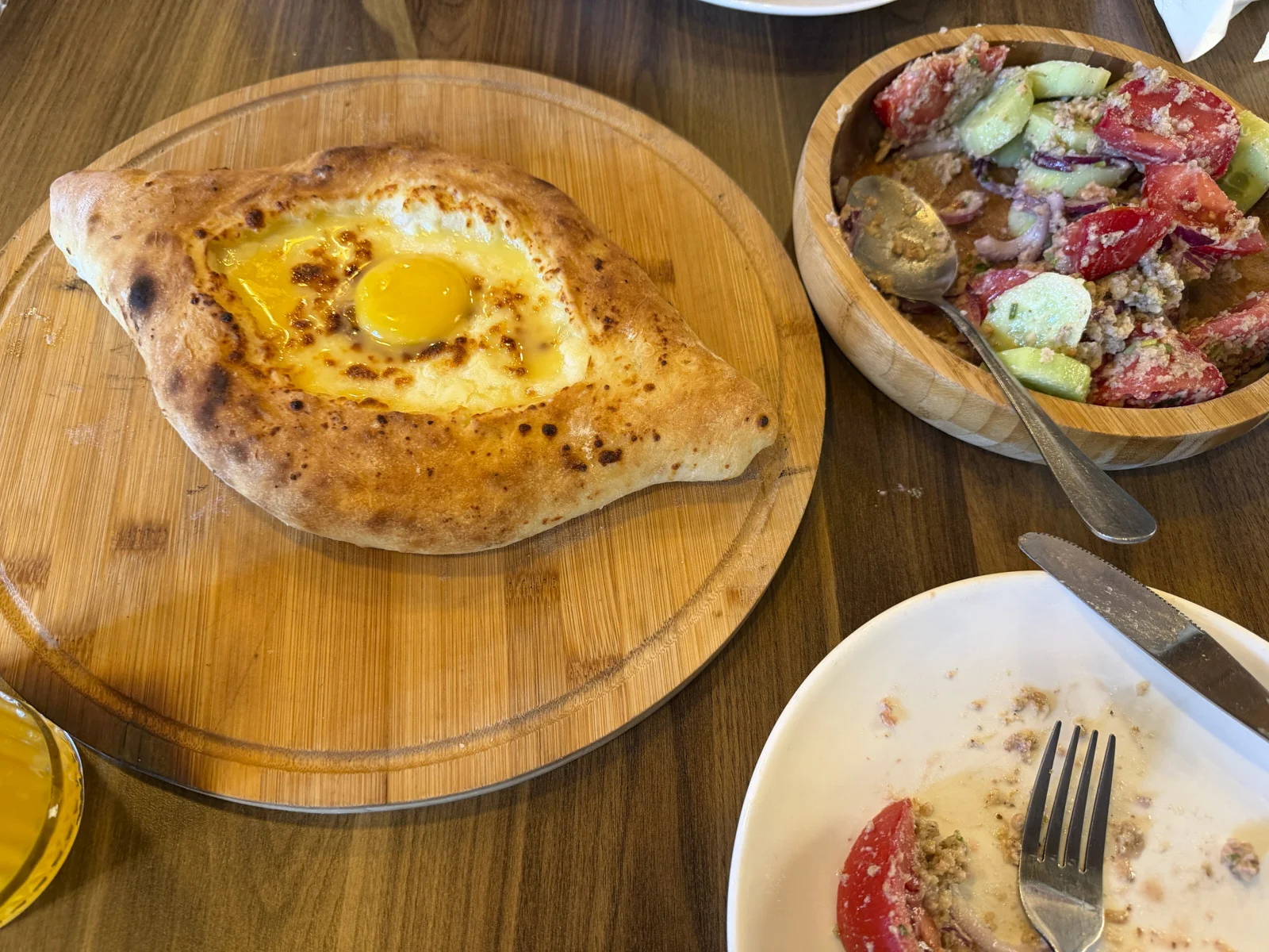 An Adjarian khachapuri with an egg, beside a bowl of tomato and cucumber salad.