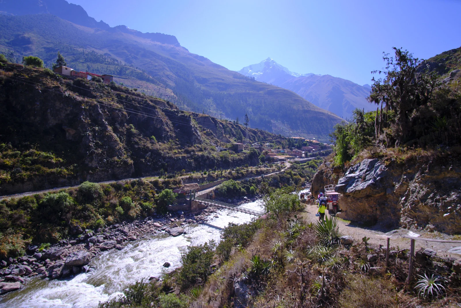 Hanging bridge over the Urubamba River at Km 82; the railway follows the far bank, and snow-capped Nevado Veronica (Wakay Willka) stands at the head of the valley