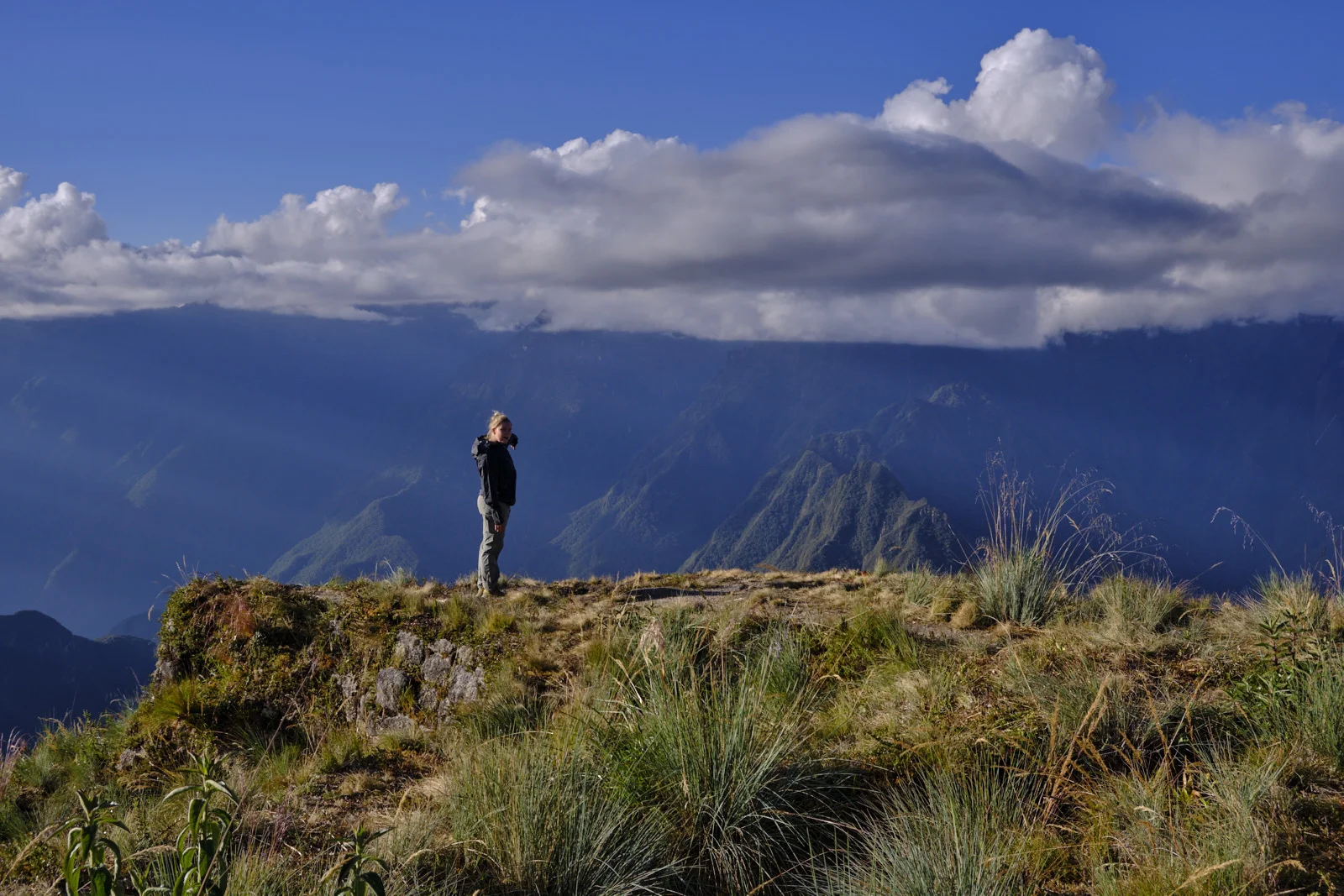 Grassy terrace edge at Phuyupatamarca looking over cloud-filled valleys and sun rays on the forested ridges